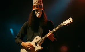 Musician wearing a white mask and KFC bucket hat plays a white electric guitar on stage.