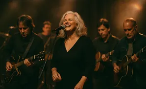 A female singer smiles and laughs into a microphone as a band plays guitars behind her.
