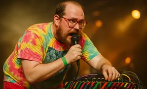 A man in a tie-dye shirt sings into a microphone, his hand adjusting knobs on a colorful audio mixer.