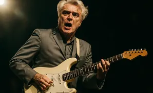 A man in a gray suit plays a light-colored electric guitar and sings into a microphone on a dark stage.