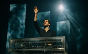 A man in a black shirt raises his hand while standing behind a clear DJ booth on a stage.