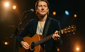 A man in a navy jacket plays an acoustic guitar on stage, with lights behind him.