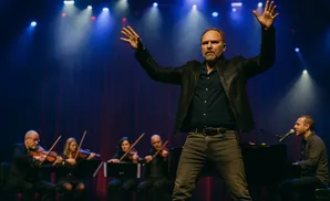 A man in a leather jacket conducting an orchestra while a pianist plays in the background.