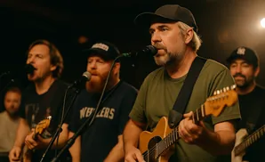 Musician in a cap sings into a microphone while playing guitar, with bandmates blurred in the background under green and orange stage lights.
