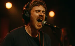A man with curly hair wearing headphones sings passionately into a microphone, with his eyes closed.