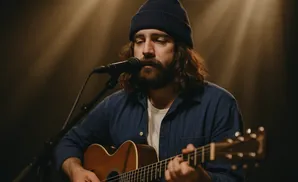Man with beard and beanie plays acoustic guitar and sings into a microphone under warm stage lighting.