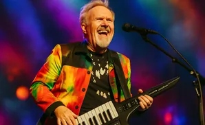 A man in a colorful jacket plays a keytar on stage, bathed in stage lights.