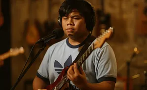 A person with headphones plays a red electric guitar in front of a microphone.