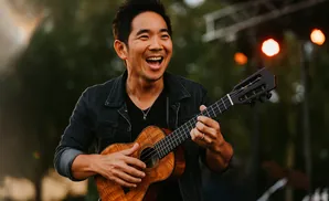 A man laughs while playing a ukulele outdoors, with lights in the background.
