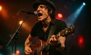 A man in a black shirt and cap sings into a microphone while playing an acoustic guitar on stage with warm lighting.
