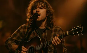 A man with long, wavy brown hair plays an acoustic guitar and sings into a microphone.