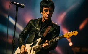 Johnny Marr plays a white Fender Jaguar guitar onstage, wearing a black leather jacket.