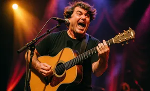 A man with messy brown hair joyfully sings and plays an acoustic guitar on stage, lit by a warm spotlight.