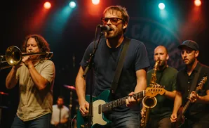 A band plays on stage. The lead singer, wearing sunglasses, plays guitar and sings into a microphone.