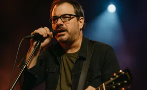 A man with glasses sings into a microphone while playing a guitar on stage.
