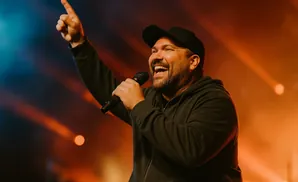 A man in a black baseball cap and hoodie smiles and points while holding a microphone on a stage with orange lighting.