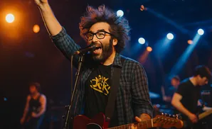 A man with glasses and a beard plays an electric guitar and sings into a microphone on a stage with bright lights.