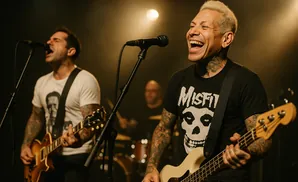 A punk band performs on stage. The lead singer, on the right, has blond hair and is laughing while playing a cream-colored bass.