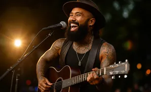 A tattooed man in a hat sings into a microphone while playing an acoustic guitar.