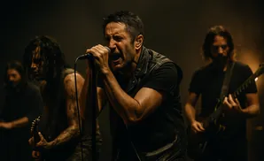 A male rock singer with a black leather vest screams into a microphone as his band performs in the background.