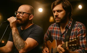Man with glasses singing into microphone next to a man playing guitar.