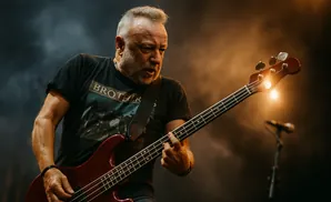 A man with a beard plays a red bass guitar on stage under warm lighting.