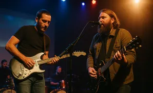 Two men play guitars on stage under colored lights. One sings into a microphone.