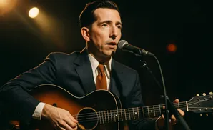 Musician playing an acoustic guitar and singing into a microphone, illuminated by stage lights.