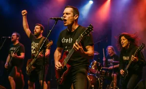 A rock band performs on stage, with a lead singer front and center playing a red guitar and singing into a microphone.