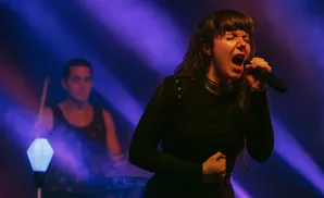 A female singer performs on stage, holding a microphone and singing with her mouth open. A drummer is visible in the background.