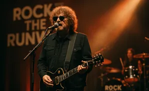 A rock guitarist with wild hair and sunglasses plays an electric guitar on stage under a spotlight.