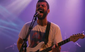 A man singing and playing a white electric guitar on stage with purple lighting.