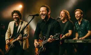 A band plays on stage, with the lead singer in the front singing into a microphone and playing a guitar.