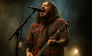 A male musician with long hair and a beard sings into a microphone while playing a red electric guitar on a dimly lit stage.