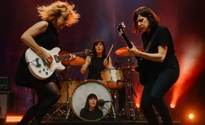 Women playing guitars and drums on stage during a live performance.