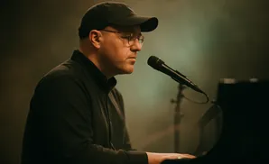 A man wearing a cap and glasses plays the piano and sings into a microphone on a dimly lit stage.