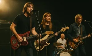 A band performs on stage with a male guitarist in front, a female bassist in the middle, and a male guitarist on the right.