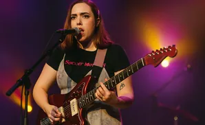 A woman with a pink hair clip sings into a microphone while playing a red electric guitar on stage.