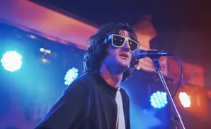 A man wearing white sunglasses sings into a microphone on a stage lit by blue and orange lights.