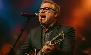 A man in a suit and glasses sings into a microphone while playing an acoustic guitar.