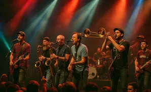 Man in a cap and striped shirt faces away from the camera on a dimly lit stage in front of an audience.