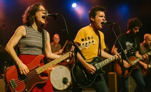 A band performs on stage, with a female bassist singing into a microphone and two guitarists behind her.