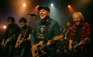 A band performs on stage with lead singer in a denim jacket and a blond guitarist in a red plaid shirt.