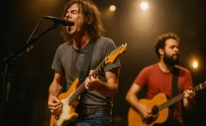 A man sings and plays an electric guitar on stage. Another man plays an acoustic guitar behind him.