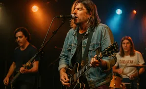 A band performs on stage with warm stage lighting, a lead singer plays guitar and sings into a microphone.