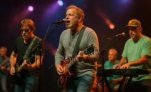 A band performs on stage, with three men playing guitars and keyboards under colorful stage lights.