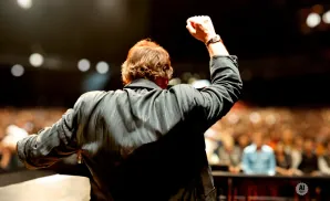 A man in a black jacket raises his fist to a cheering crowd from a stage.