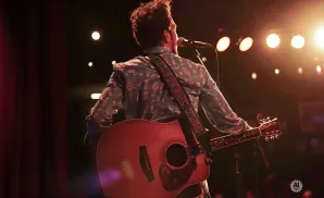 A musician plays an acoustic guitar on stage, illuminated by warm stage lights.