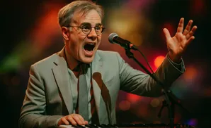 Man in a light blue suit and tie singing into a microphone at a keyboard, with his hand raised.