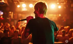 A singer with short blonde hair and tattoos performs into a microphone on a stage, with a blurred crowd in the background.